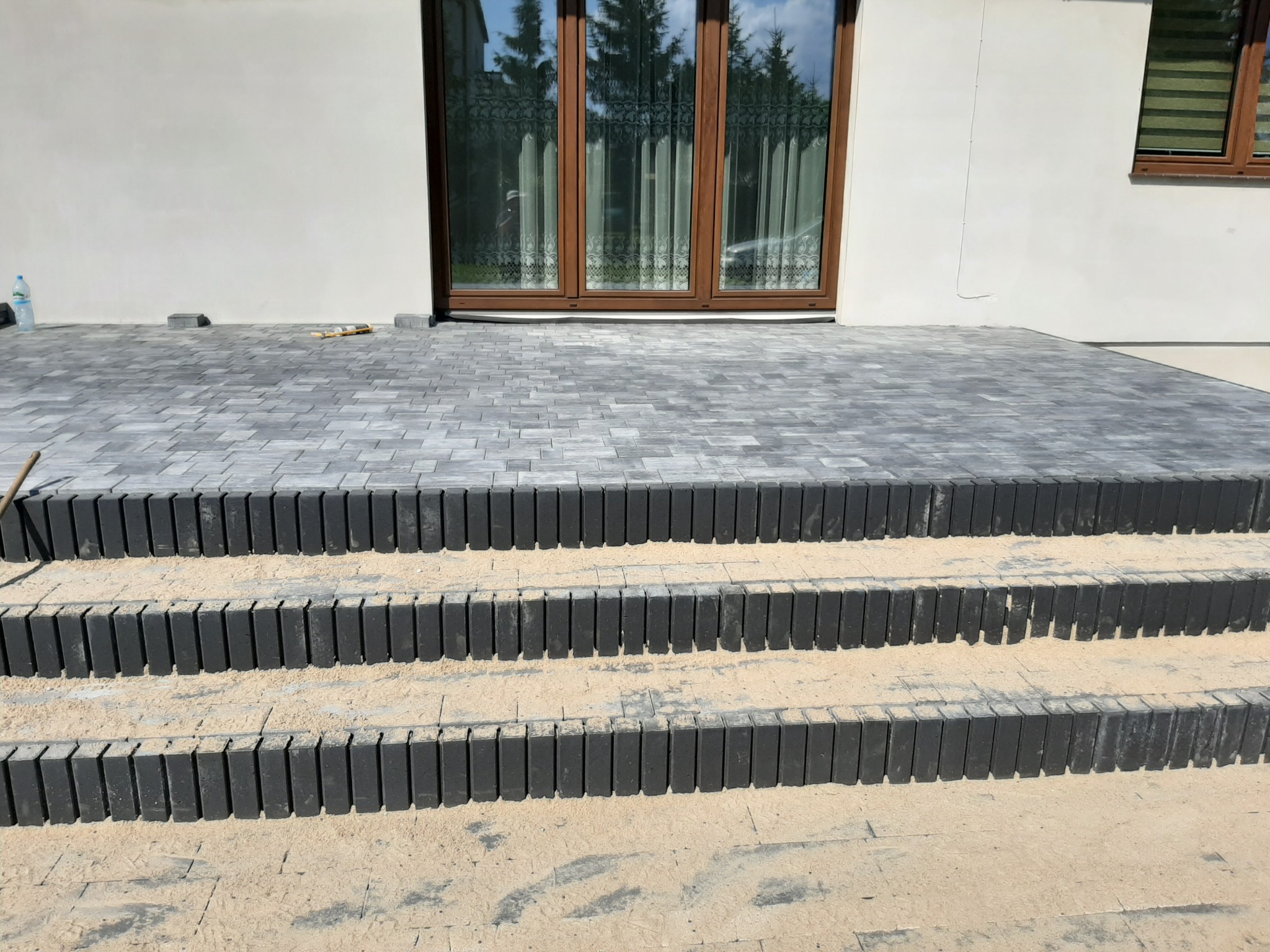 Schody zewnętrzne z kostki brukowej w kolorze szarym, prowadzące do drzwi wejściowych domu. Widoczne trzy stopnie z czarnych, pionowo ułożonych elementów oddzielających stopnie. Na stopniach piasek.