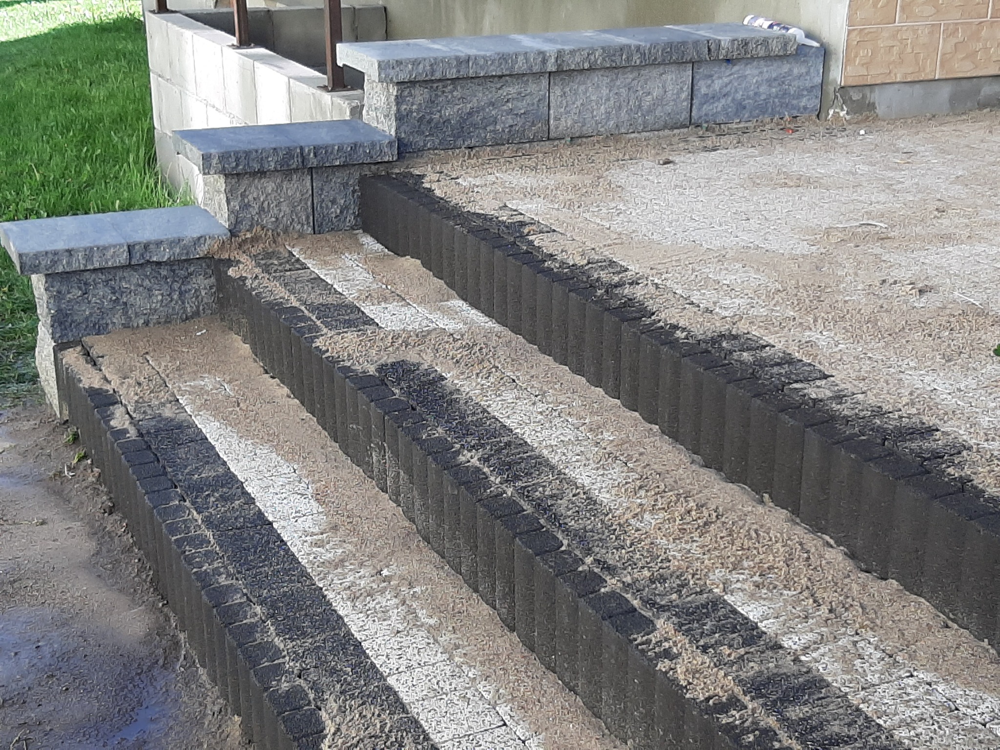 Schody zewnętrzne w trakcie budowy, z widocznymi stopniami z kostki brukowej i elementami wykończeniowymi z kamienia, zasypane piaskiem.