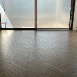 Ułożona w jodełkę, brązowa podłoga z płytek ceramicznych w jasnym, nowym pomieszczeniu z dużymi oknami zabezpieczonymi folią ochronną.