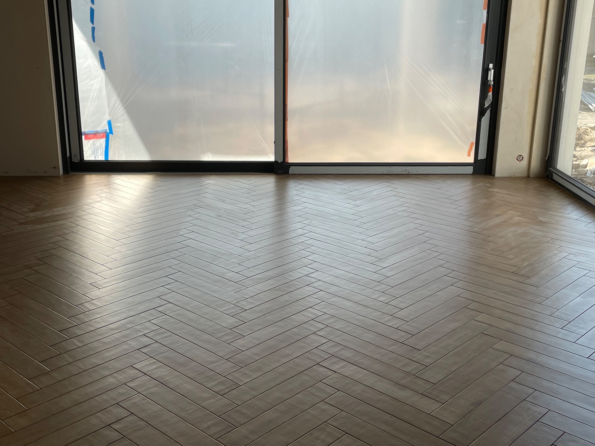 Ułożona w jodełkę, brązowa podłoga z płytek ceramicznych w jasnym, nowym pomieszczeniu z dużymi oknami zabezpieczonymi folią ochronną.