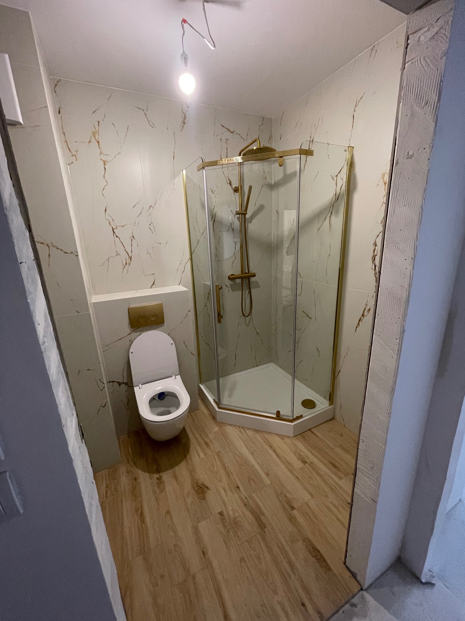 Wykończona łazienka z kabiną prysznicową w złotych ramach, wiszącą toaletą i marmurowymi płytkami na ścianach, oświetlona pojedynczą żarówką.