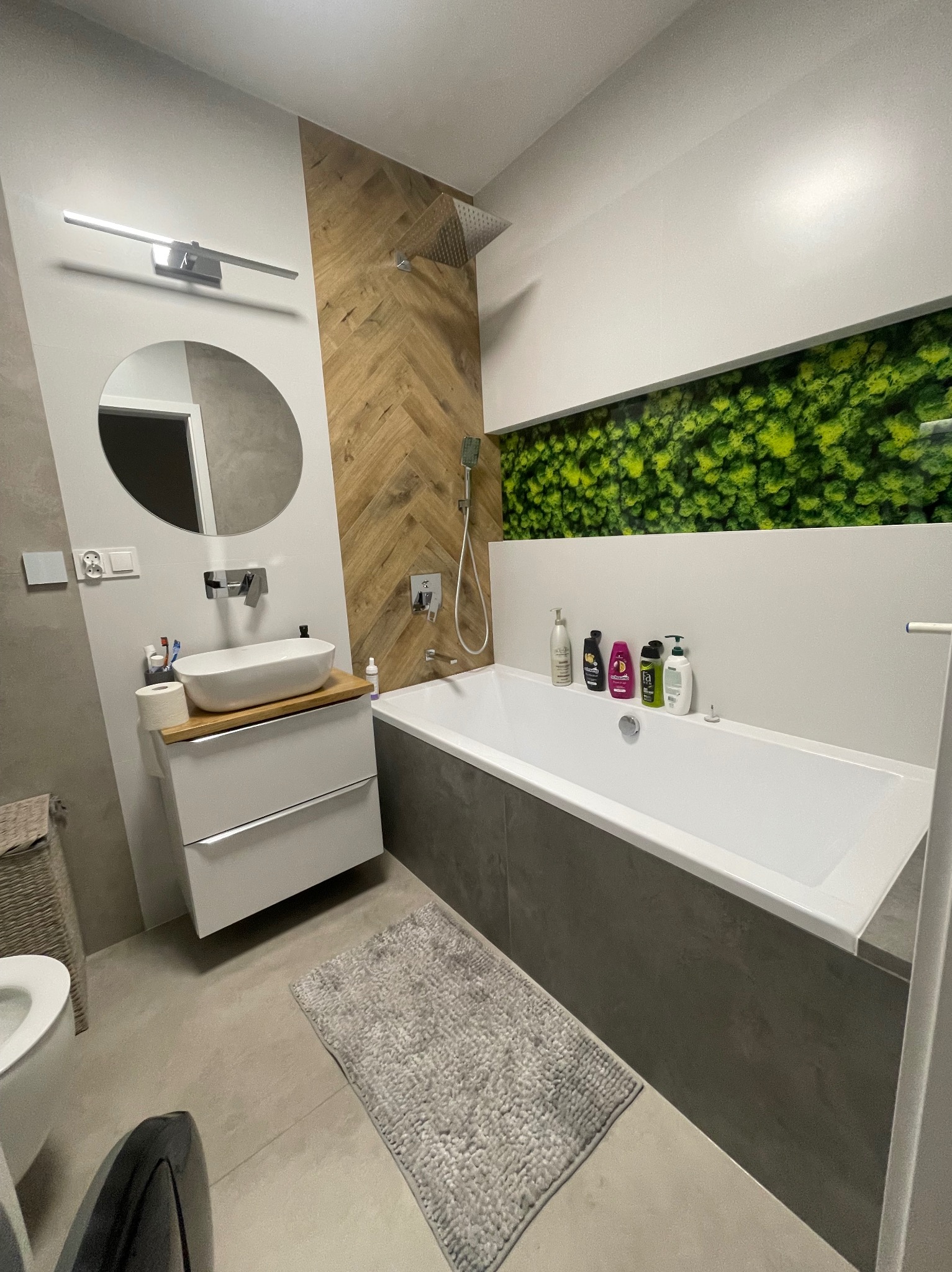 Nowoczesna łazienka z drewnianą ścianą ułożoną w jodełkę, okrągłym lustrem nad umywalką, wanną obudowaną szarą płytką i panelem z zielenią. Na wannie ustawione kosmetyki w różnych opakowaniach.