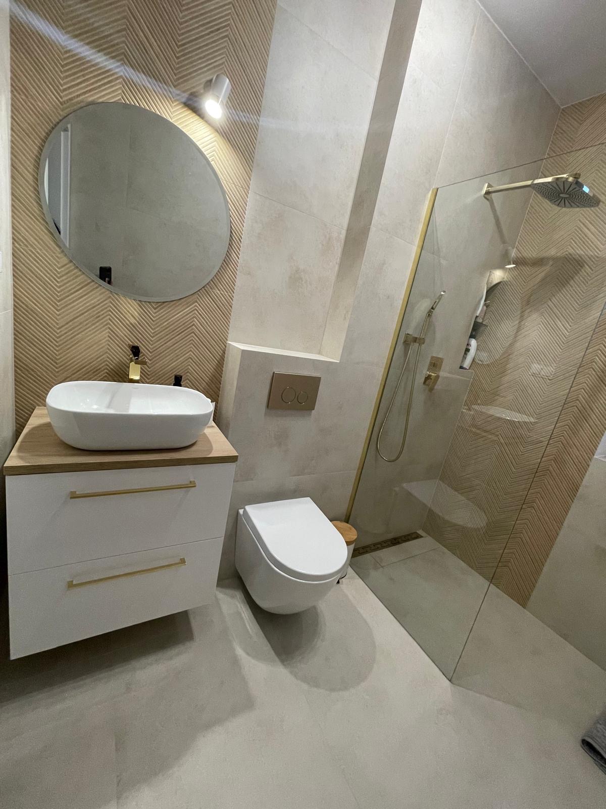 Nowoczesna łazienka z kabiną prysznicową typu walk-in, umywalką nablatową na szafce z szufladami z złotymi uchwytami, wiszącą miską WC i geometrycznymi wzorami na ścianach.