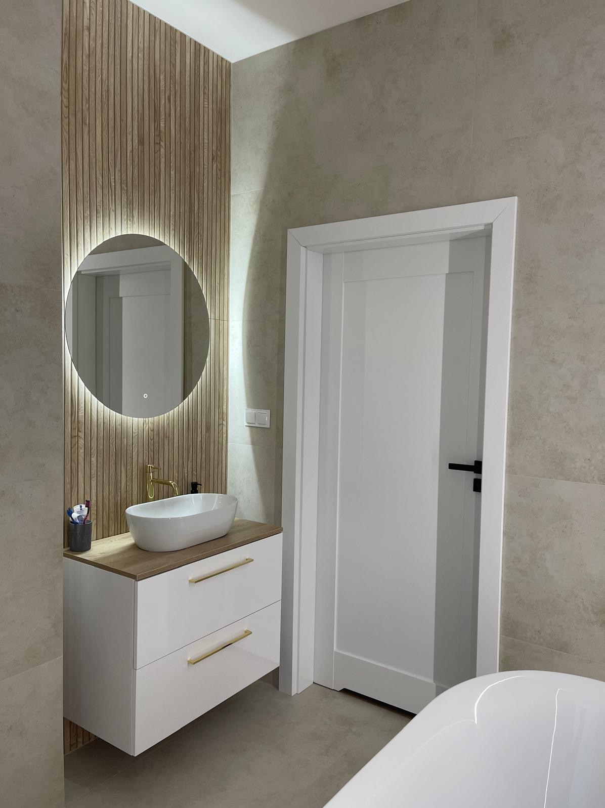 Nowoczesna łazienka z umywalką nablatową, okrągłym lustrem z podświetleniem LED, drewnianą ścianką dekoracyjną i białą szafką z złotymi uchwytami.
