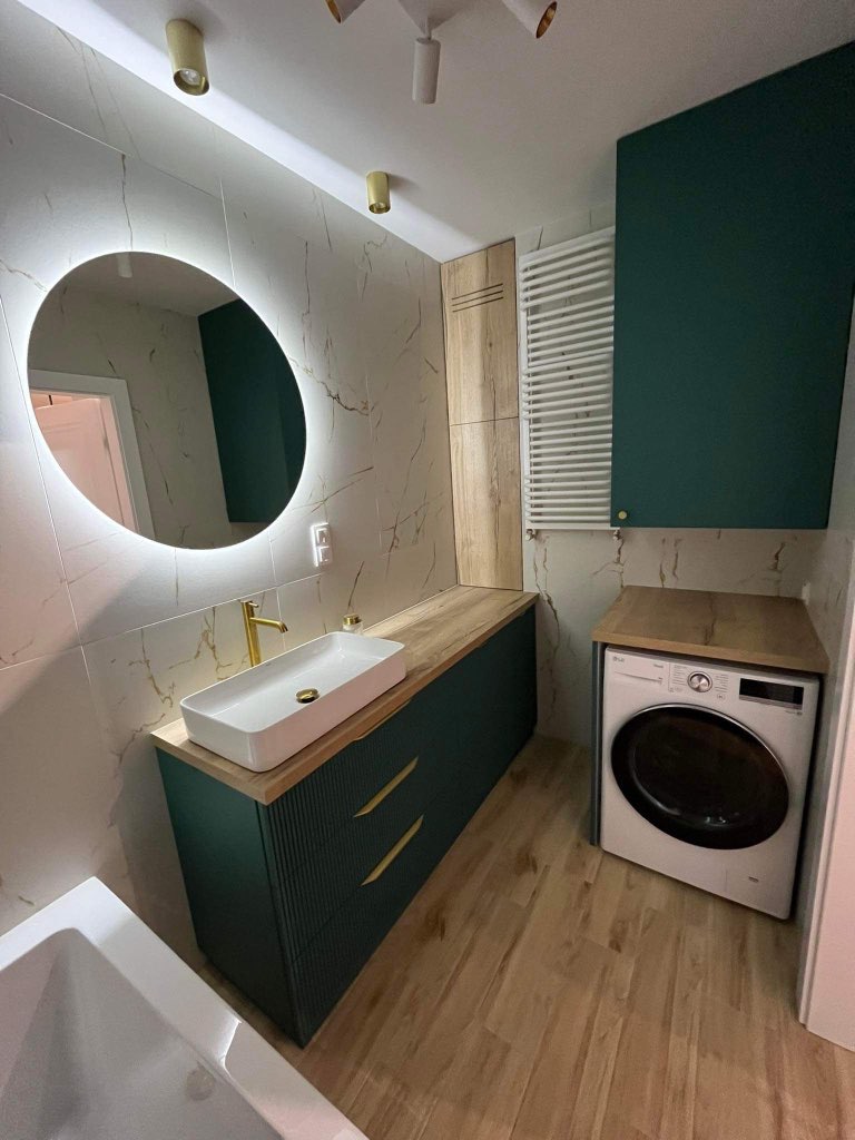 Nowoczesna łazienka z umywalką nablatową, szafką w kolorze butelkowej zieleni z frezowanymi frontami i złotymi uchwytami, okrągłym lustrem z podświetleniem LED, pralką pod blatem i grzejnikiem...
