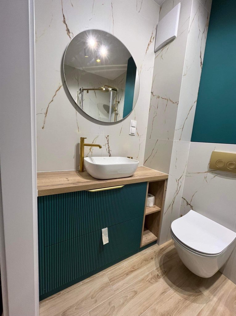 Nowoczesna łazienka z umywalką nablatową, armaturą w kolorze złotym i szafką w odcieniach zieleni. W tle widoczne płytki imitujące marmur oraz kabina prysznicowa.