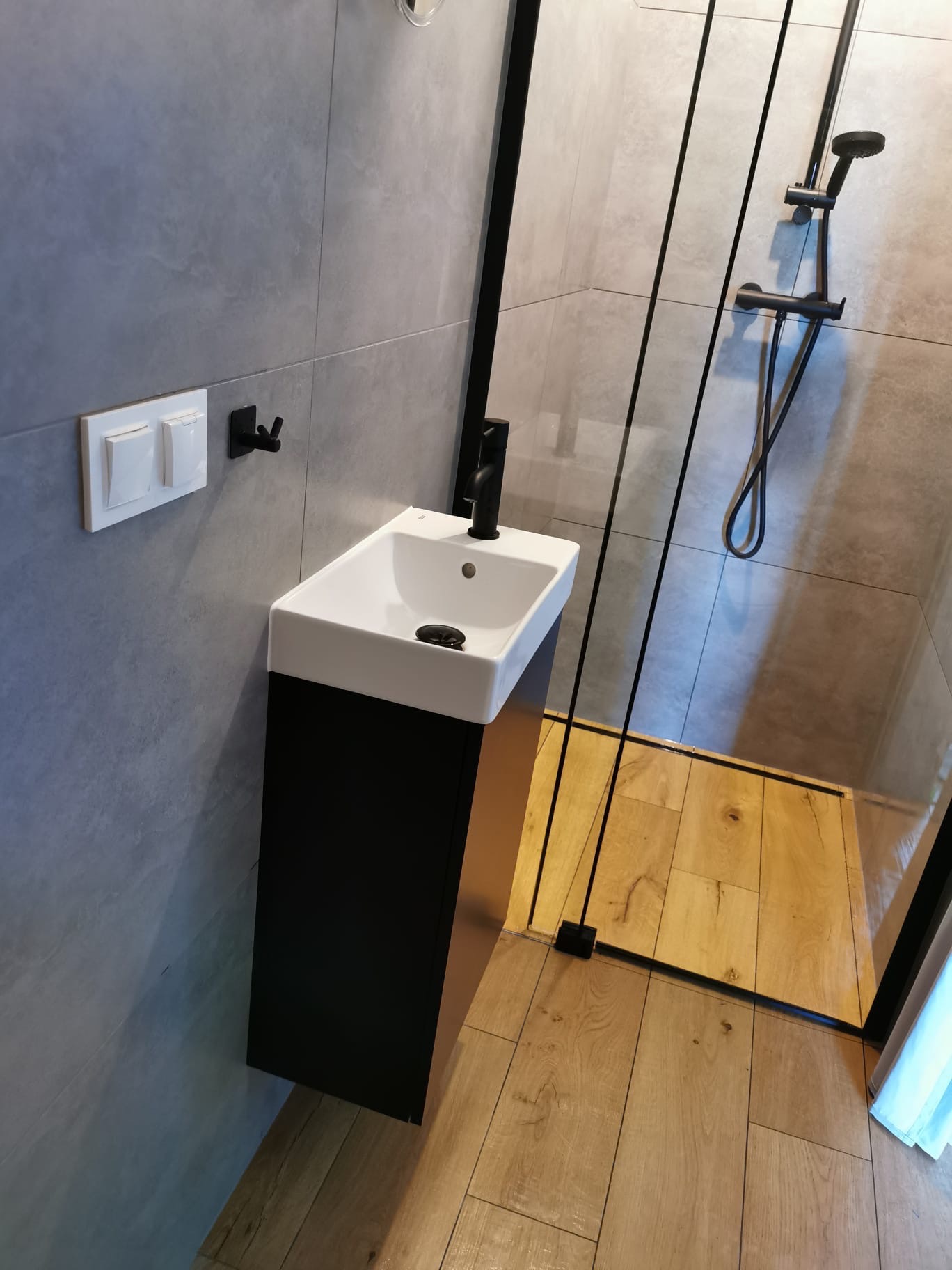 Nowoczesna łazienka z szarą płytką imitującą kamień, wisząca biała umywalka z czarną armaturą, kabina prysznicowa z czarnymi profilami i drewnopodobną podłogą, minimalistyczny włącznik światła...