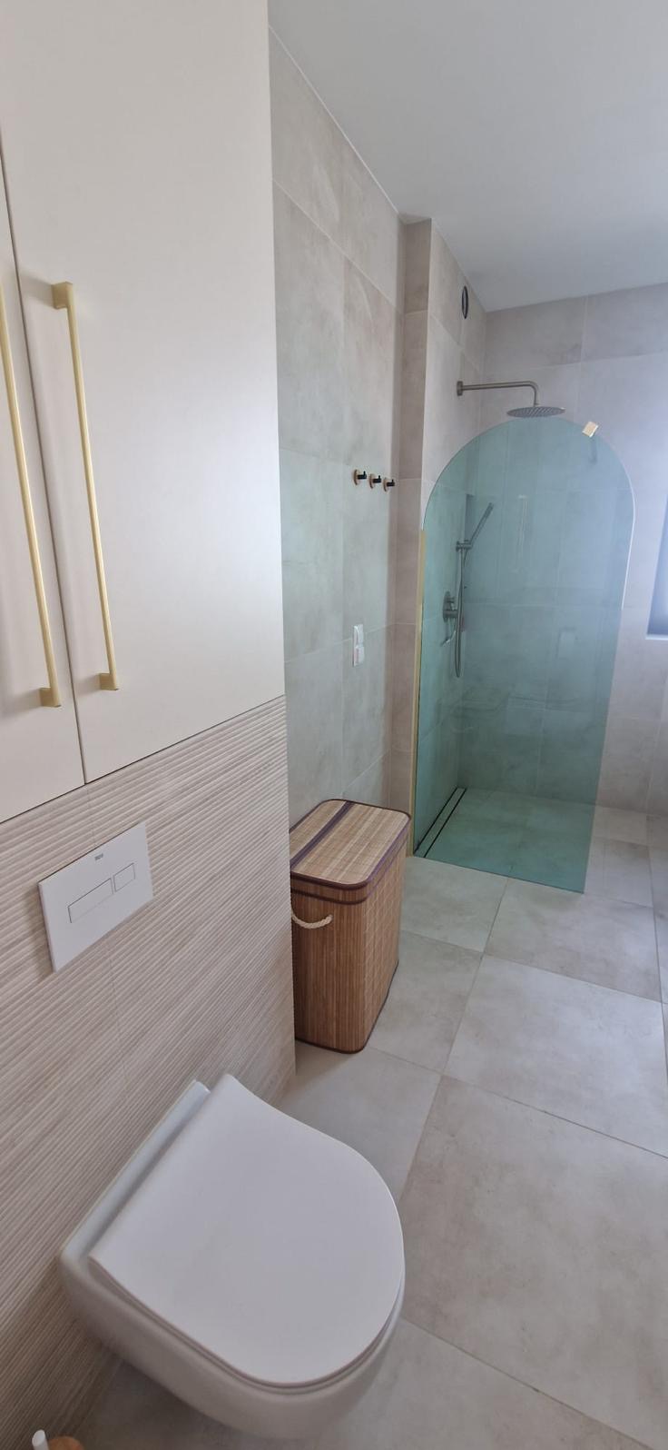 Jasna łazienka z kabiną prysznicową o zaokrąglonym kształcie, wiszącą toaletą i bambusowym koszem na pranie, wykończona płytkami w odcieniach beżu.