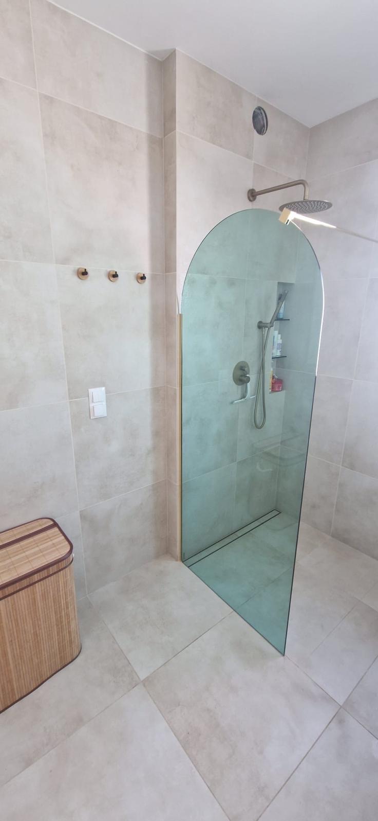 Beżowa łazienka z kabiną prysznicową typu walk-in z zaokrąglonym szkłem, odpływem liniowym i trzema drewnianymi wieszakami.