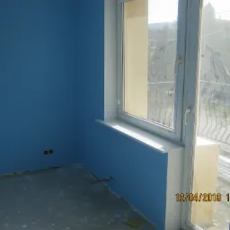 Pokój w trakcie malowania na niebiesko, widoczne niedoskonałości na podłodze i ścianach, brudne okno z widokiem na zewnątrz, wystające kable ze ściany.