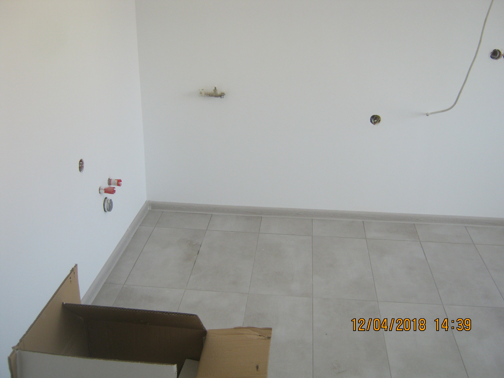 Fragment wnętrza w trakcie wykańczania: szare płytki na podłodze, białe ściany z wystającymi przewodami i rurkami, kartonowe pudło w rogu.