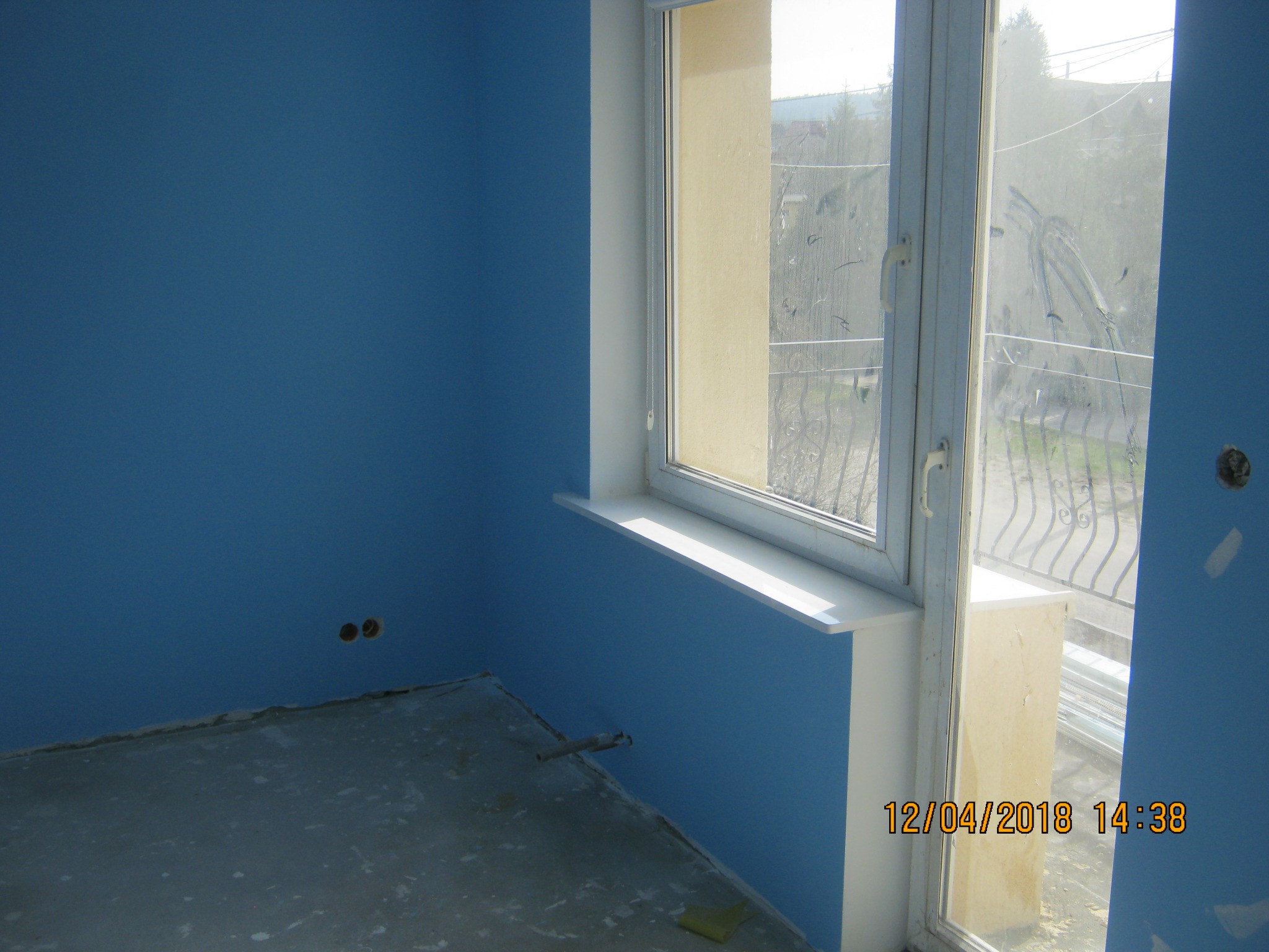 Pokój w trakcie malowania na niebiesko, widoczne niedoskonałości na podłodze i ścianach, brudne okno z widokiem na zewnątrz, wystające kable ze ściany.