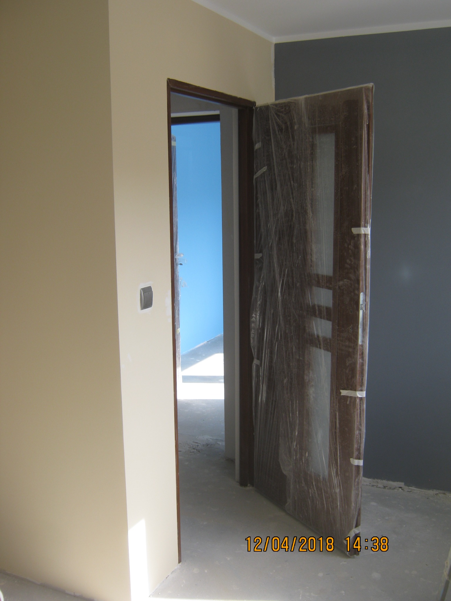 Nowe drewniane drzwi wewnętrzne, zabezpieczone folią ochronną, w trakcie montażu w nowym pomieszczeniu z pomalowanymi ścianami w odcieniach beżu i szarości. Widoczny fragment ściany...