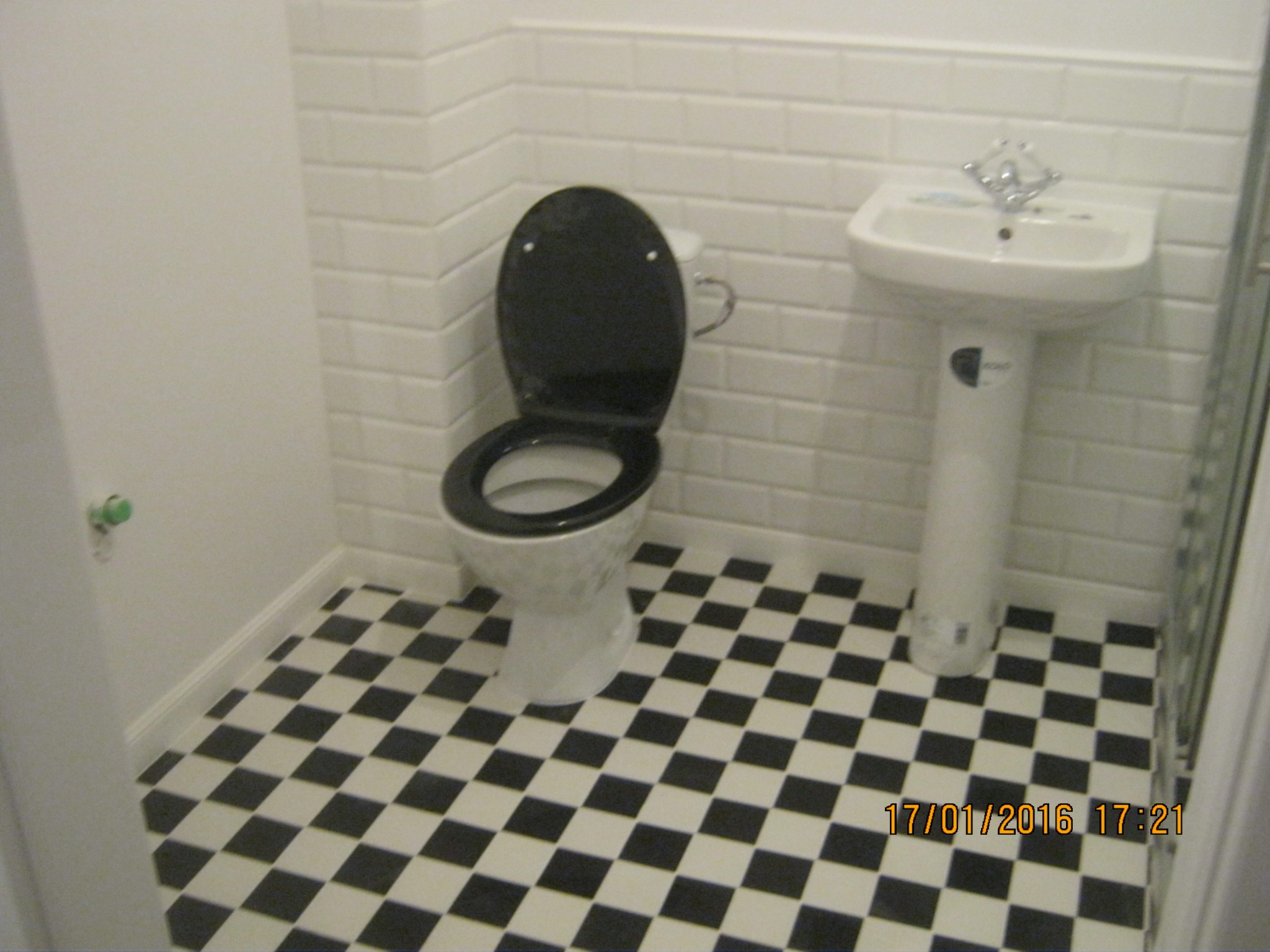 Biała toaleta i umywalka na postumencie w łazience z podłogą w czarno-białą szachownicę i ścianami wyłożonymi białymi płytkami imitującymi cegłę.