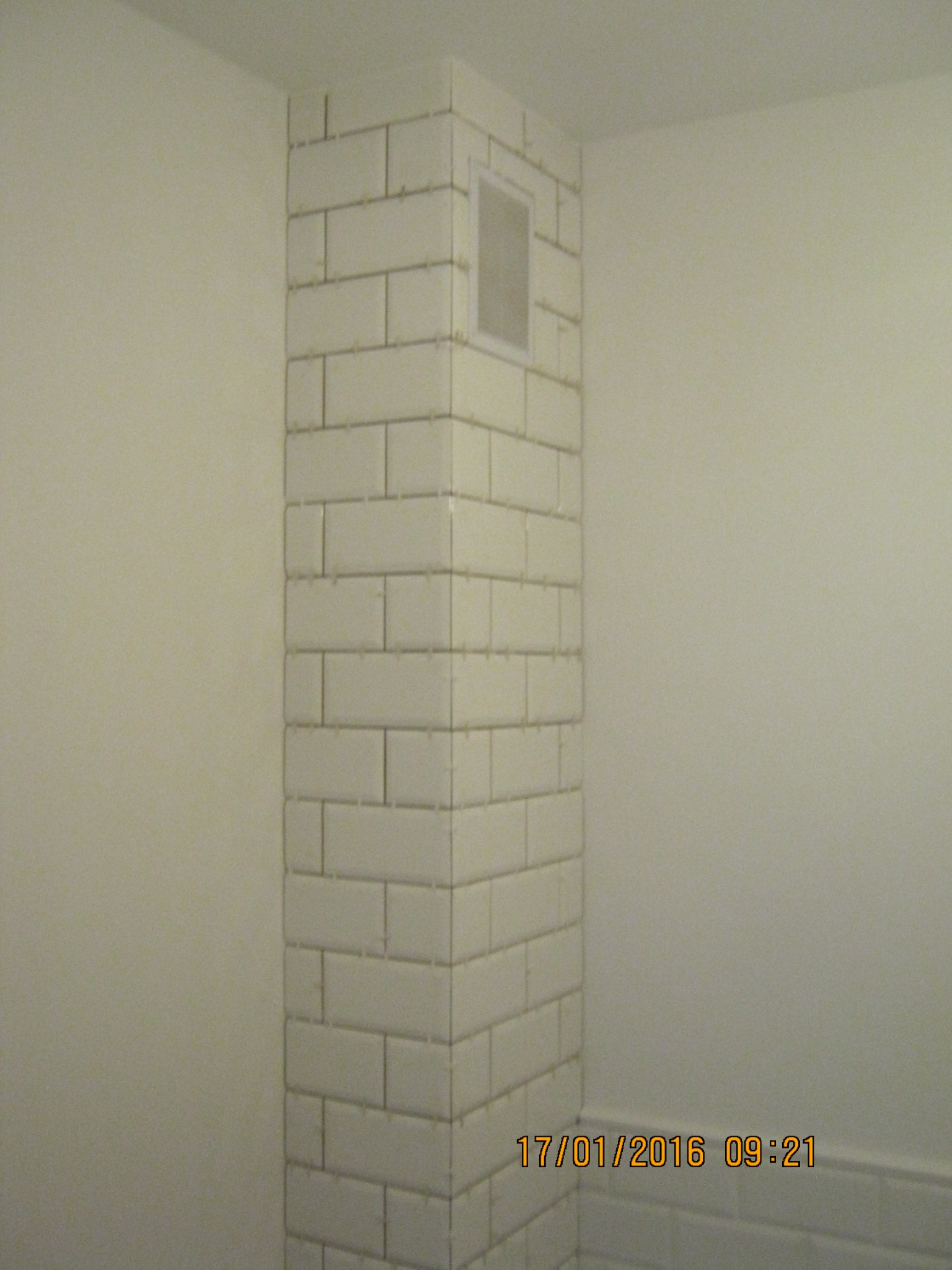 Obudowa pionu wentylacyjnego wykończona białymi, prostokątnymi płytkami ceramicznymi z widocznymi krzyżykami dystansowymi, na tle pomalowanej na kremowo ściany.