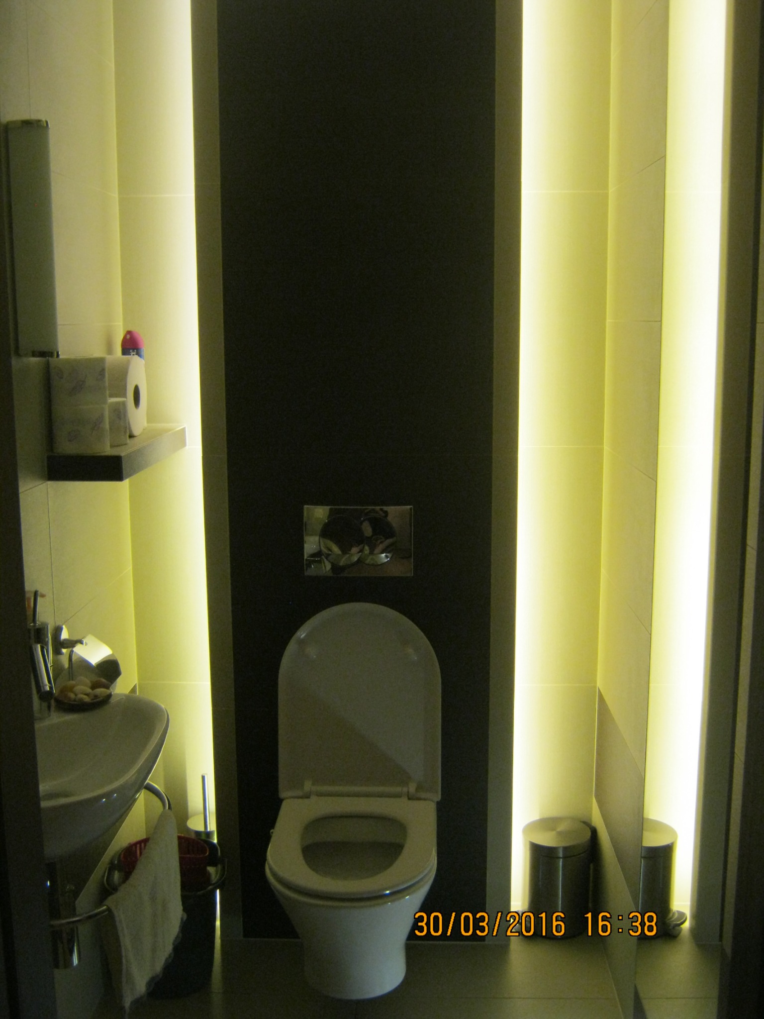 Nowoczesna toaleta z podświetleniem LED w pionowych wnękach, umywalka z baterią i półką na papier toaletowy, uchwycona z perspektywy drzwi.