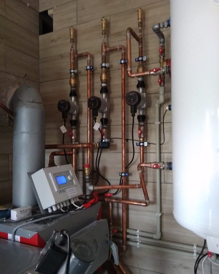 Kompleksowa instalacja grzewcza z rur miedzianych, widoczne pompy obiegowe, sterownik z niebieskim wyświetlaczem, biały zasobnik ciepłej wody, zamontowane na ścianie z płytek imitujących drewno.