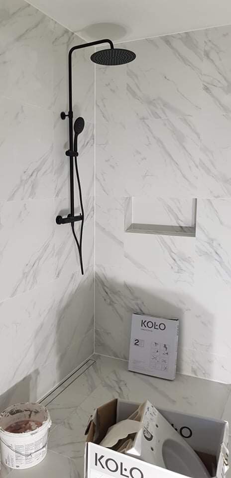 Nowoczesna kabina prysznicowa z czarną armaturą i marmurowymi płytkami, widoczne puste opakowania po ceramice Koło i wiadro z zaprawą.