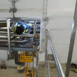 Spawacz w kombinezonie i masce ochronnej, pracujący na wysokości na skomplikowanym systemie rur w hali przemysłowej, widoczna platforma robocza Ramirent i instalacja kablowa.