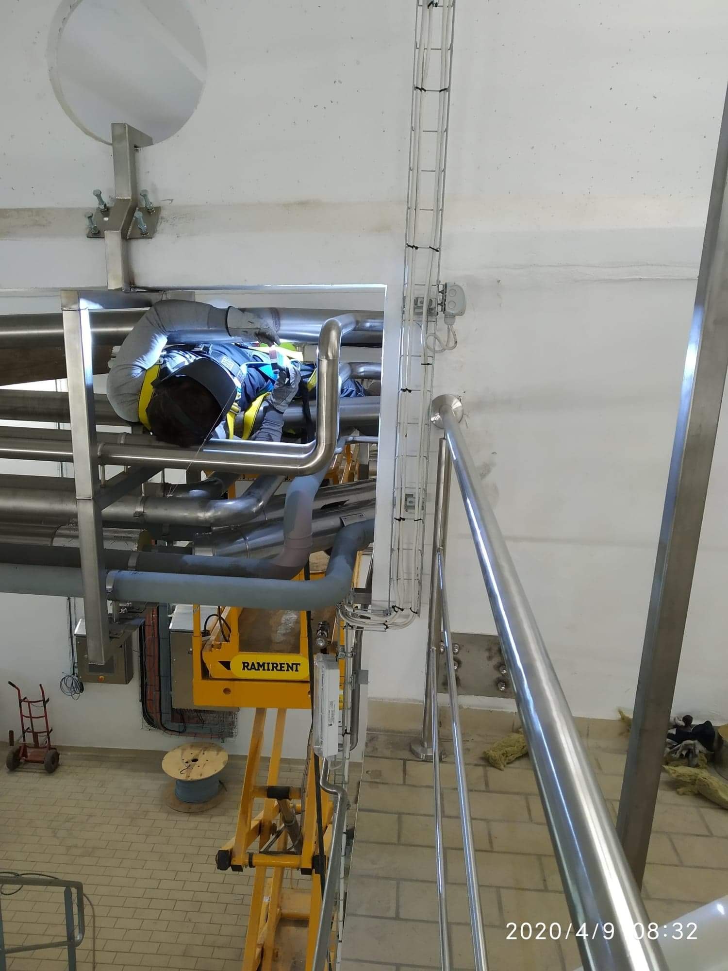 Spawacz w kombinezonie i masce ochronnej, pracujący na wysokości na skomplikowanym systemie rur w hali przemysłowej, widoczna platforma robocza Ramirent i instalacja kablowa.