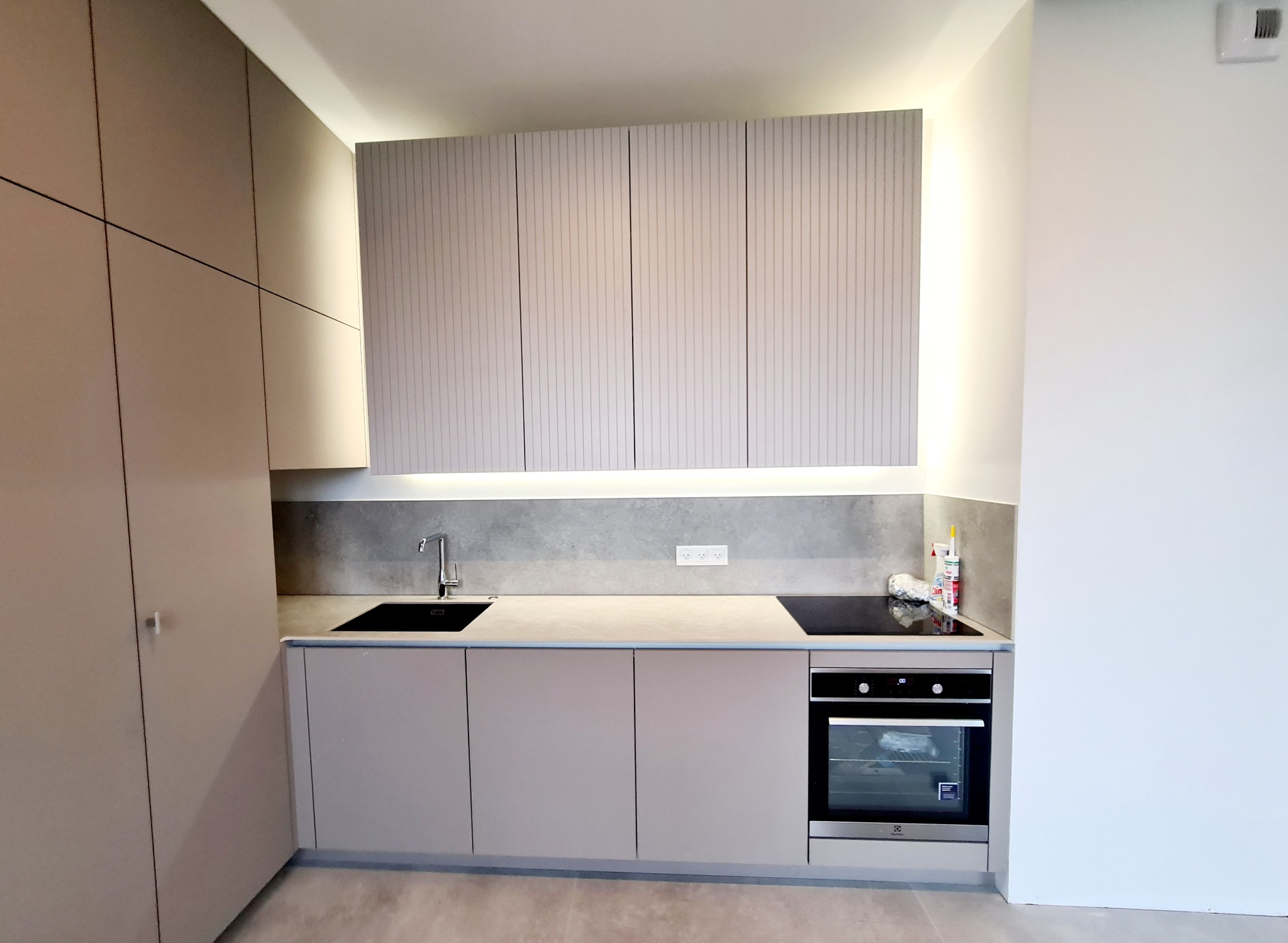 Nowoczesna kuchnia z beżowymi szafkami, podświetleniem LED pod górnymi szafkami i szarym blatem, z czarnym zlewem i płytą indukcyjną.