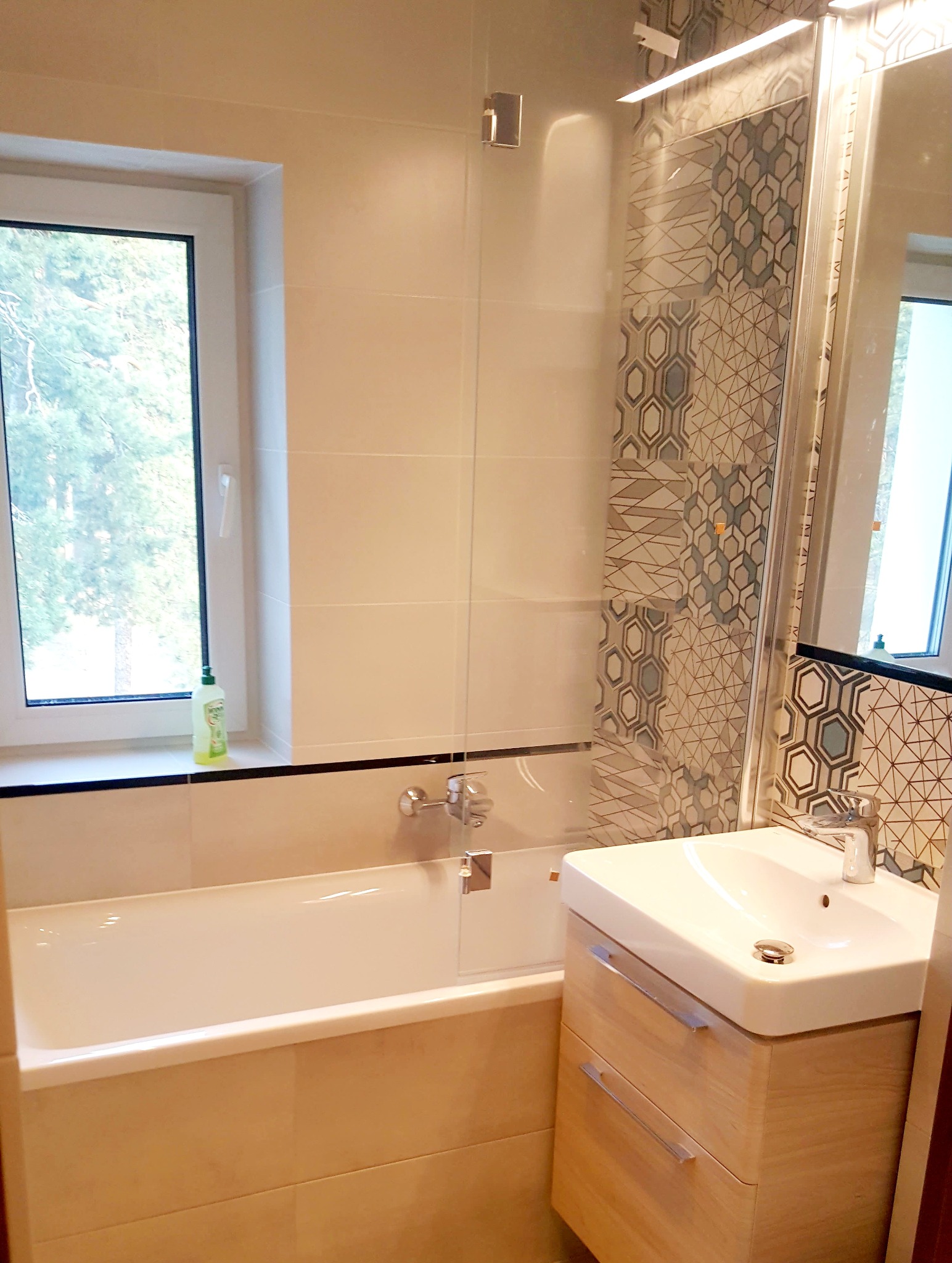 Jasna łazienka po remoncie z wanną, umywalką z szafką z jasnego drewna oraz geometrycznymi płytkami na ścianie pod prysznicem.