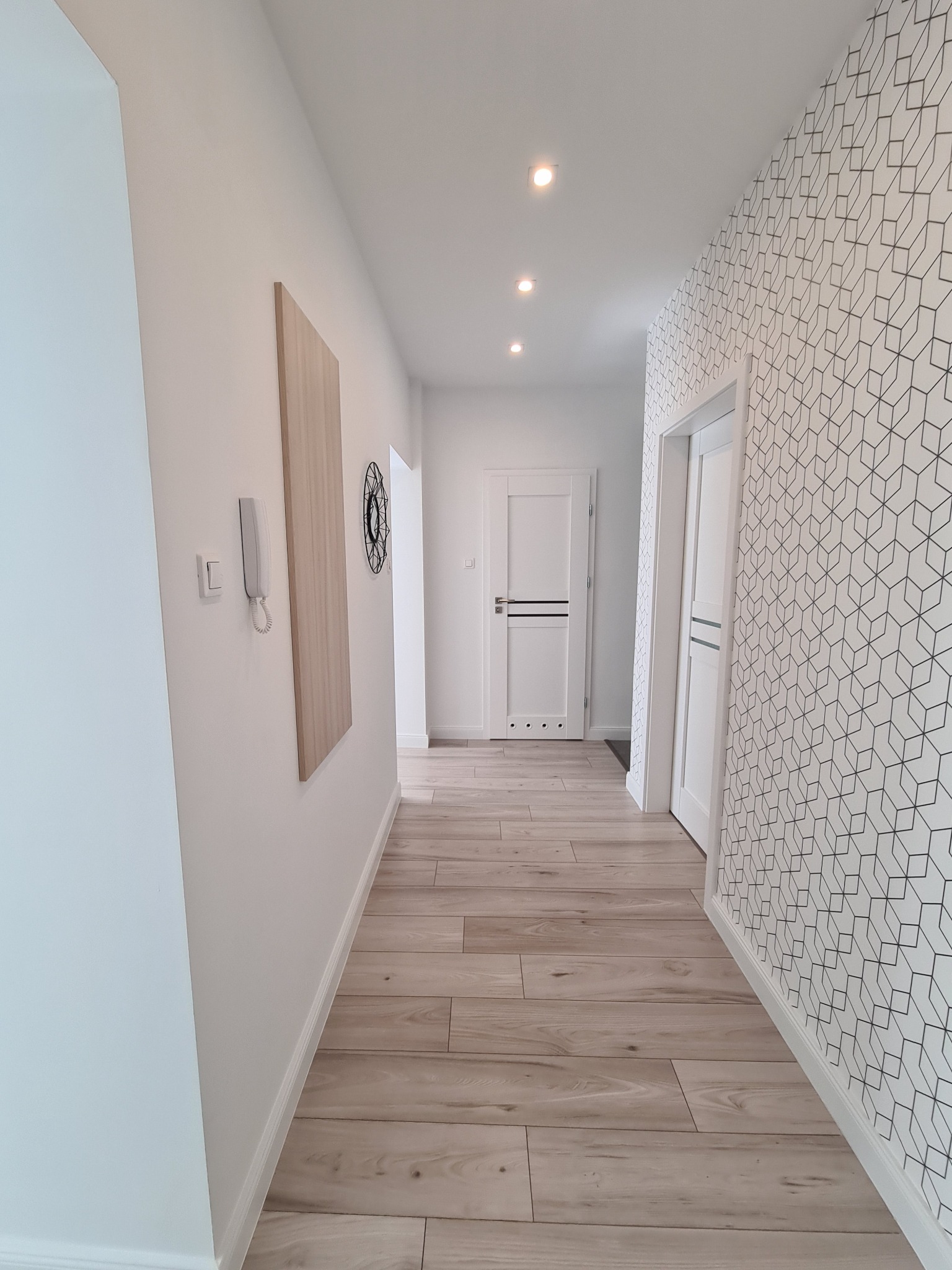 Nowoczesny przedpokój z panelami podłogowymi imitującymi drewno, białą ścianą z interkomem i drewnianym panelem dekoracyjnym, oraz ścianą z geometryczną tapetą w czarno-białe romby.