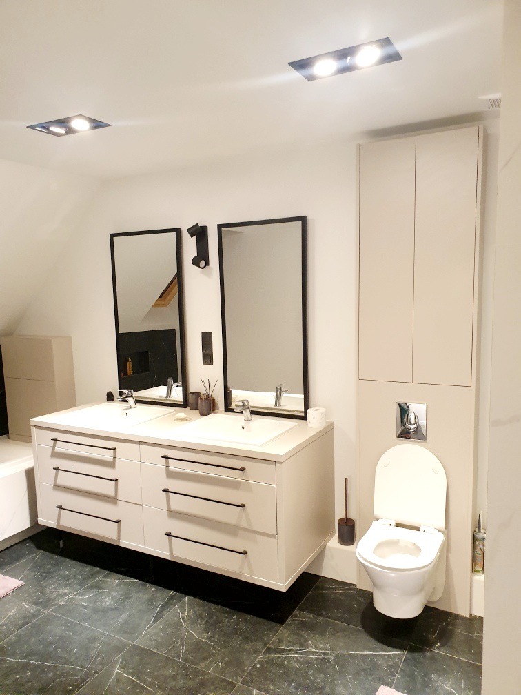 Jasna łazienka z podwójną umywalką na szafce z ciemnymi uchwytami, toaleta w zabudowie, duże lustra w czarnych ramach i ciemna marmurowa podłoga.