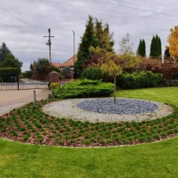 Uporządkowany ogród przydomowy z centralnym okręgiem wysypanym szarymi kamieniami, otoczonym kostką brukową i świeżo posadzoną trawą, z młodym drzewkiem w środku, widok na bramę i dom w tle.