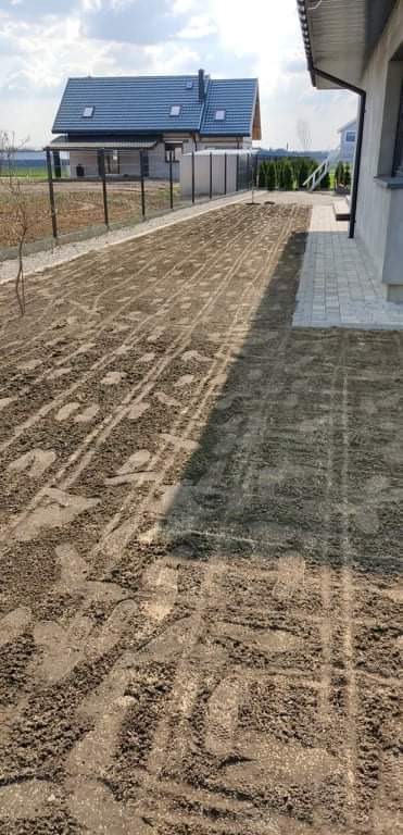 Przygotowany teren pod trawnik z widocznymi śladami po bronowaniu, obok kostka brukowa i dom z ciemnym dachem.