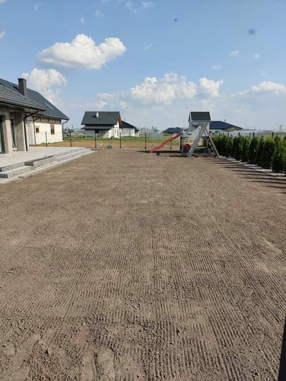Przygotowany teren pod trawnik z widocznymi śladami po bronowaniu, w tle domy jednorodzinne i plac zabaw z czerwoną zjeżdżalnią.