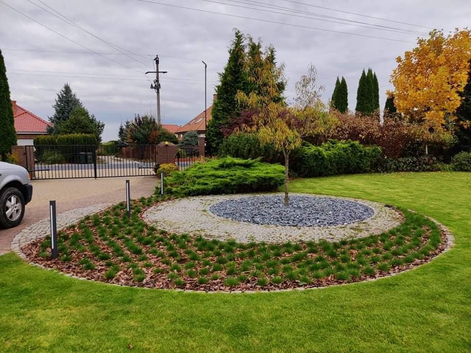 Uporządkowany ogród przydomowy z centralnym okręgiem wysypanym szarymi kamieniami, otoczonym kostką brukową i świeżo posadzoną trawą, z młodym drzewkiem w środku, widok na bramę i dom w tle.