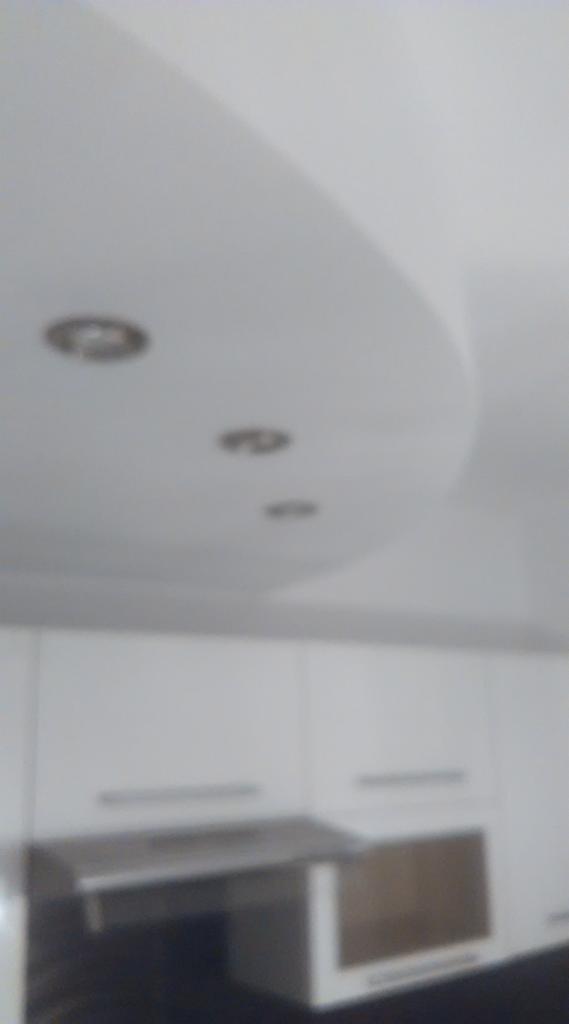 Fragment sufitu z trzema okrągłymi lampami halogenowymi wpuszczonymi w podwieszany, gipsowy sufit o zaokrąglonym kształcie, poniżej widoczne białe szafki kuchenne i fragment okapu.