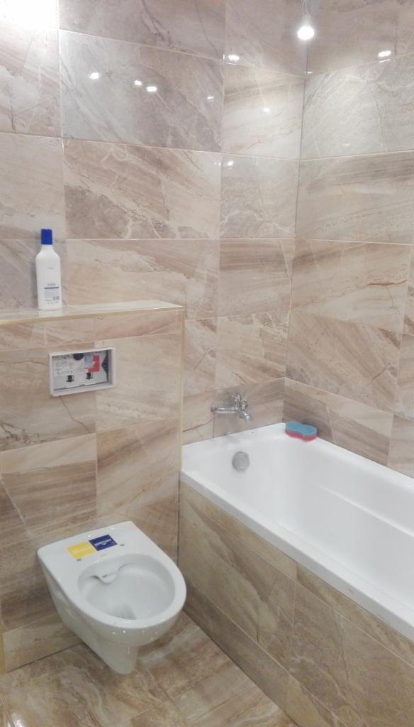 Wykończona łazienka z beżowymi płytkami imitującymi marmur, wisząca toaleta z żółtą naklejką producenta, biała wanna z chromowaną baterią i niebiesko-czerwoną gąbką, biała butelka z niebieską...