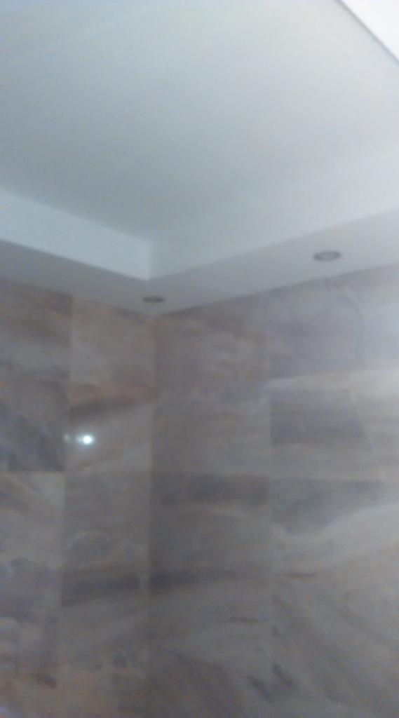 Ściana wyłożona płytkami imitującymi marmur w odcieniach szarości i beżu, z widocznymi dwoma punktowymi lampami sufitowymi.