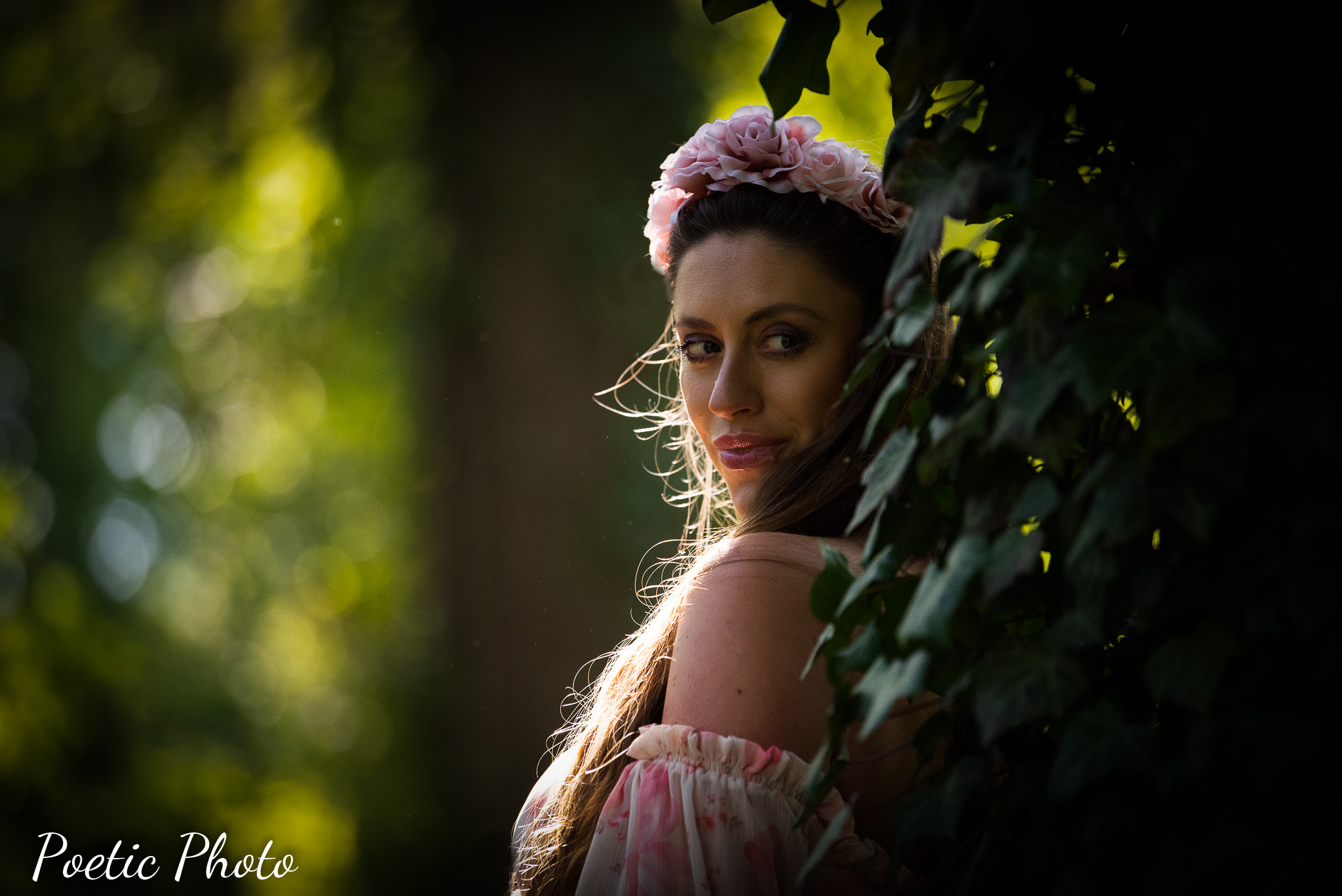 Portret kobiety z wiankiem z różowych kwiatów we włosach, spoglądającej przez liście bluszczu, z delikatnym oświetleniem od tyłu.