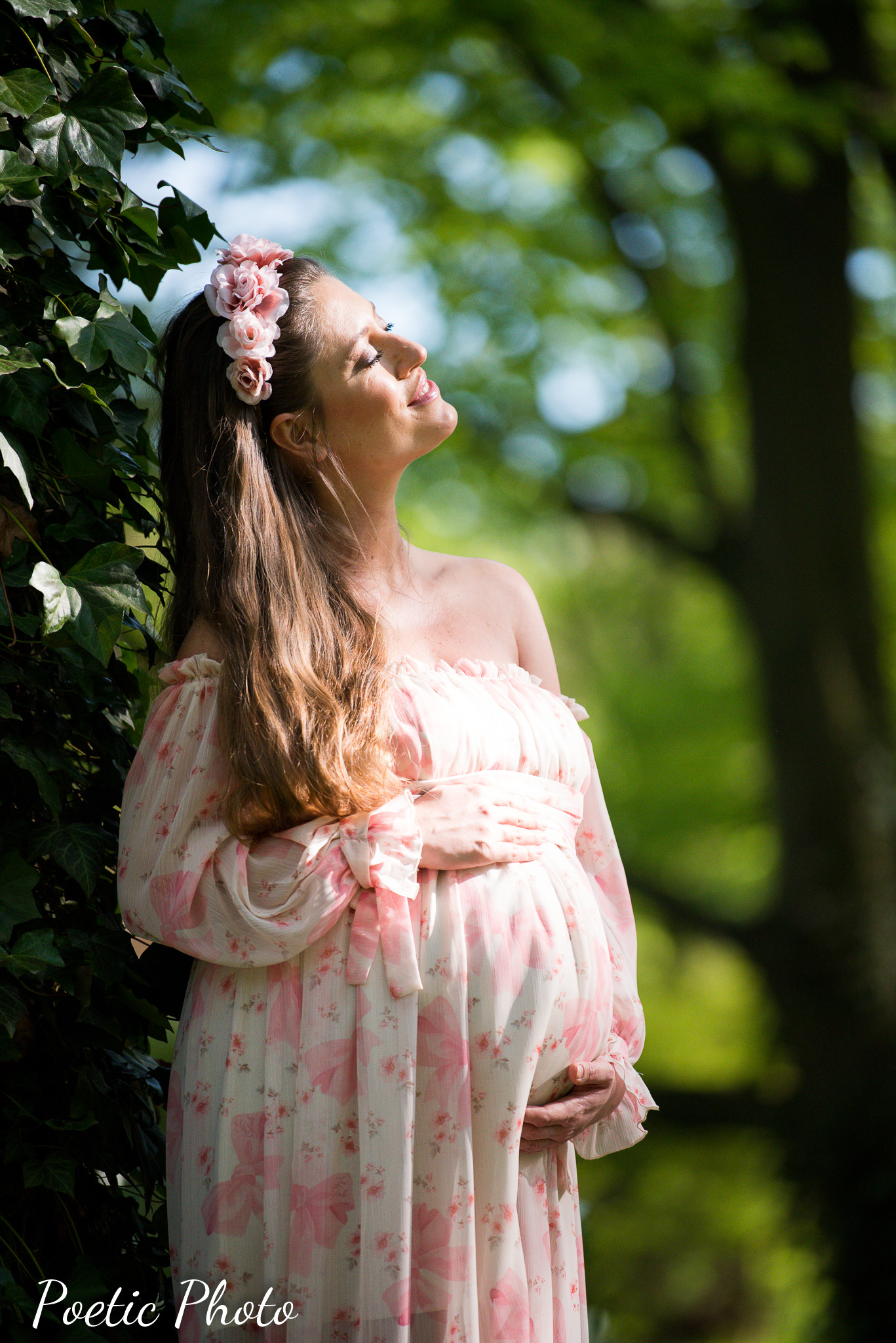 Kobieta w ciąży w zwiewnej sukience w kwiaty, z wiankiem z różowych kwiatów we włosach, opiera się o drzewo, z głową uniesioną ku słońcu.