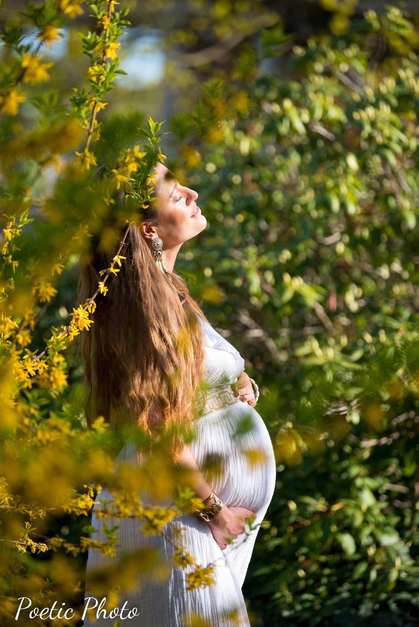 Kobieta w ciąży w zwiewnej białej sukience, z profilu, otoczona kwitnącymi gałązkami forsycji, z delikatnym makijażem i złotą biżuterią, spogląda w górę w promieniach słońca.