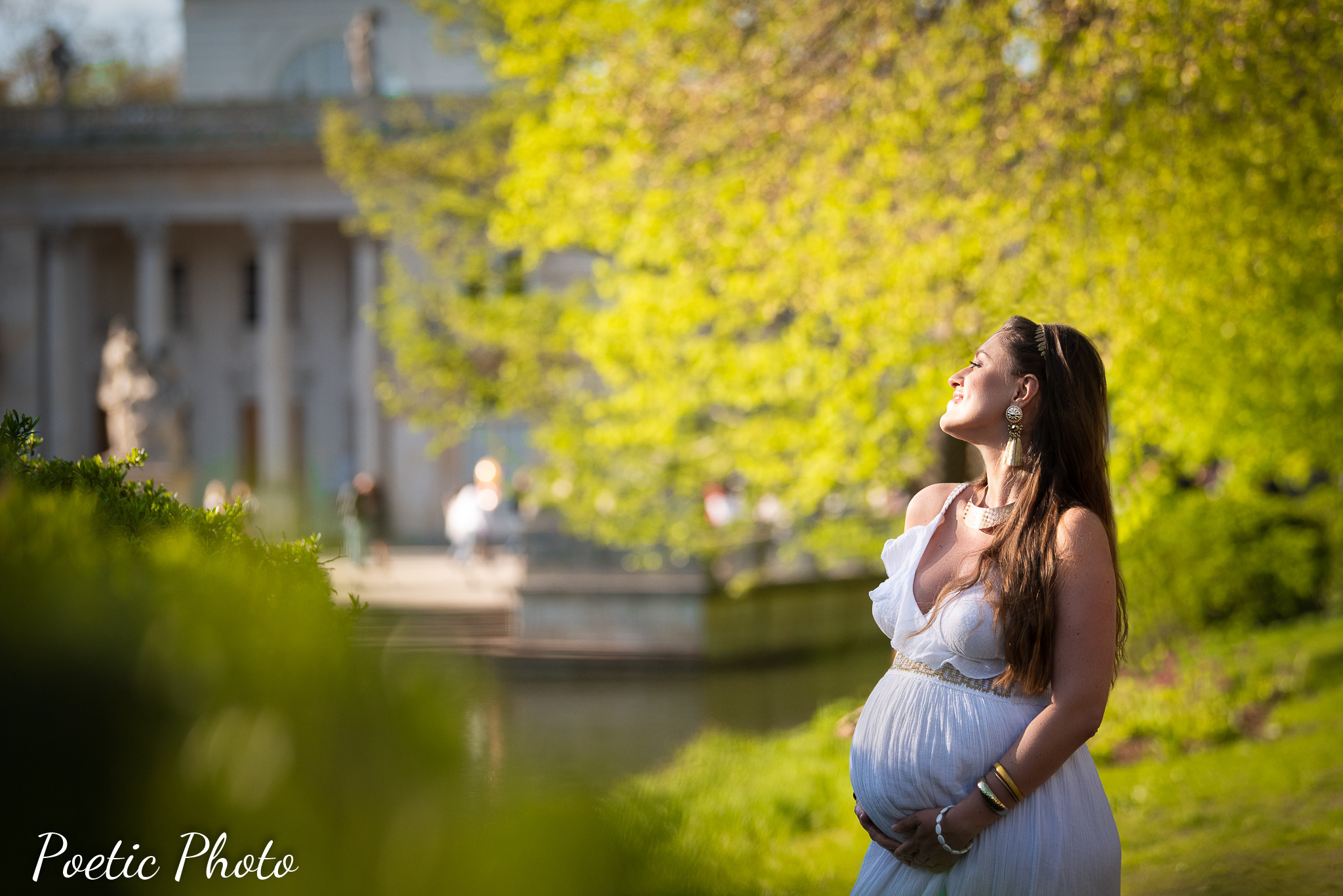 Uśmiechnięta kobieta w białej sukni ciążowej, z rękami na brzuchu, pozuje na tle parku z klasycystyczną architekturą w tle, oświetlona ciepłym, słonecznym światłem.