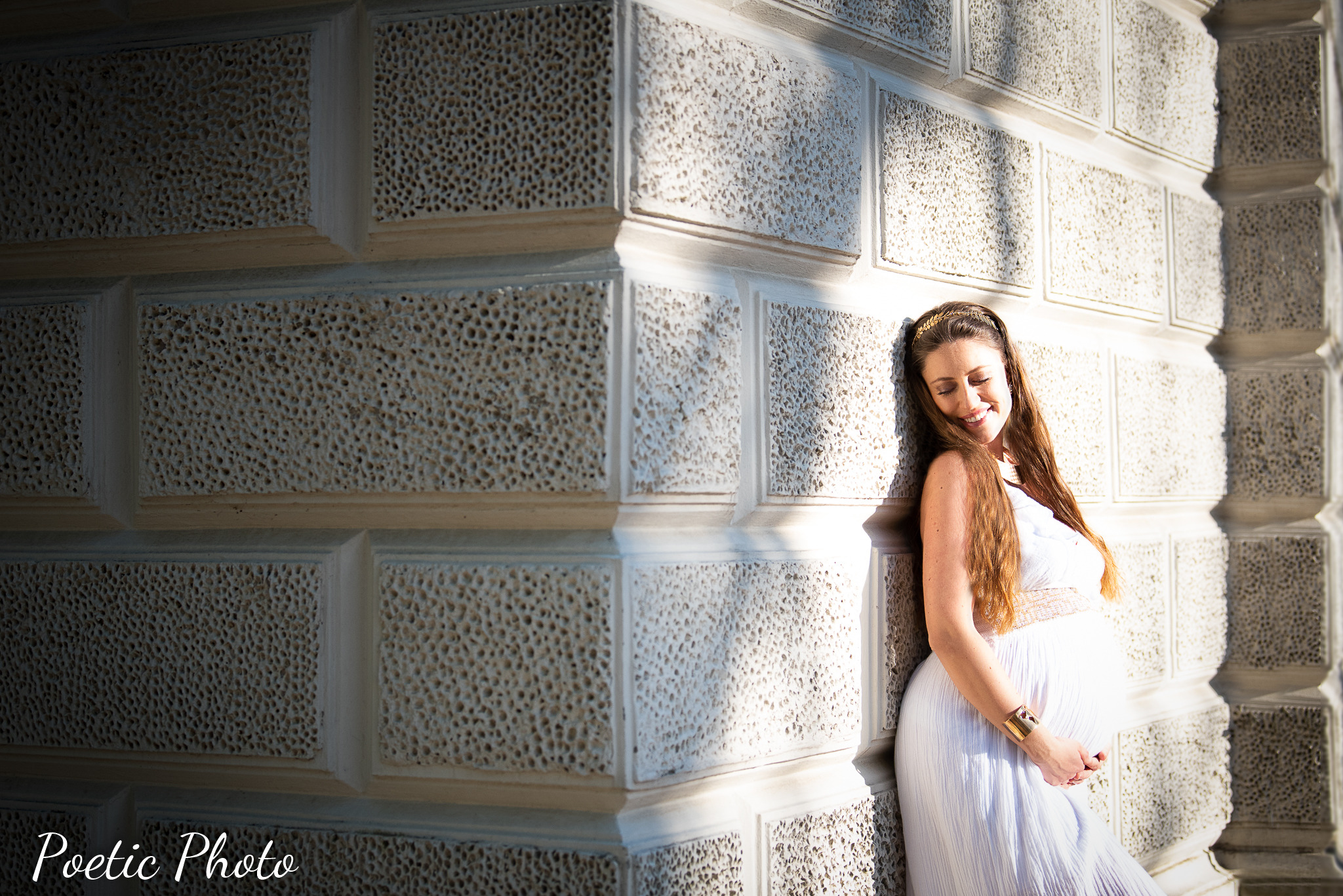 Uśmiechnięta kobieta w białej sukni ciążowej, oparta o narożną ścianę z porowatego kamienia w promieniach słońca.