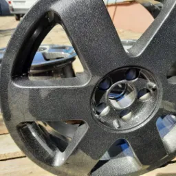 Błyszcząca, ciemnoszara felga aluminiowa z widoczną strukturą lakieru, leżąca na drewnianych deskach w słoneczny dzień. Druga felga w tle. Widoczne otwory montażowe i ramiona felgi.