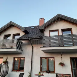 Dwupiętrowy dom z dwoma balkonami z poziomymi, szarymi barierkami, udekorowany wiszącymi światełkami, z oknami z brązowymi ramami i ciemnym dachem