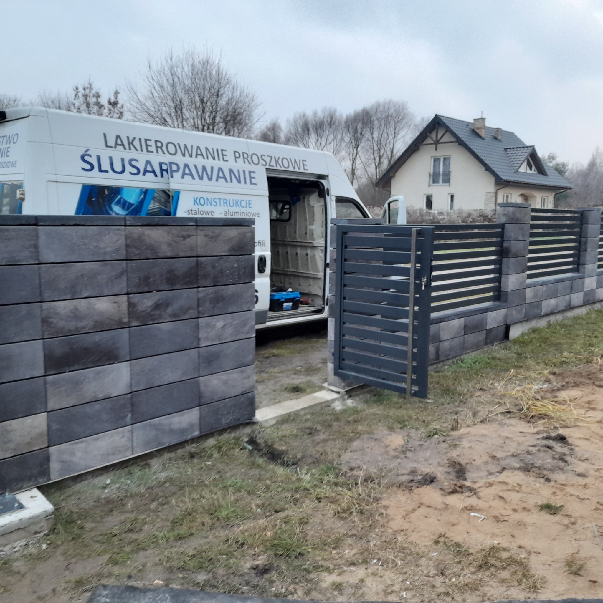 Nowoczesna brama wjazdowa z poziomymi panelami i ogrodzenie z szarych bloczków betonowych, częściowo zasłonięte przez otwarte drzwi białego samochodu dostawczego firmy 'Lakierowanie Proszkowe...