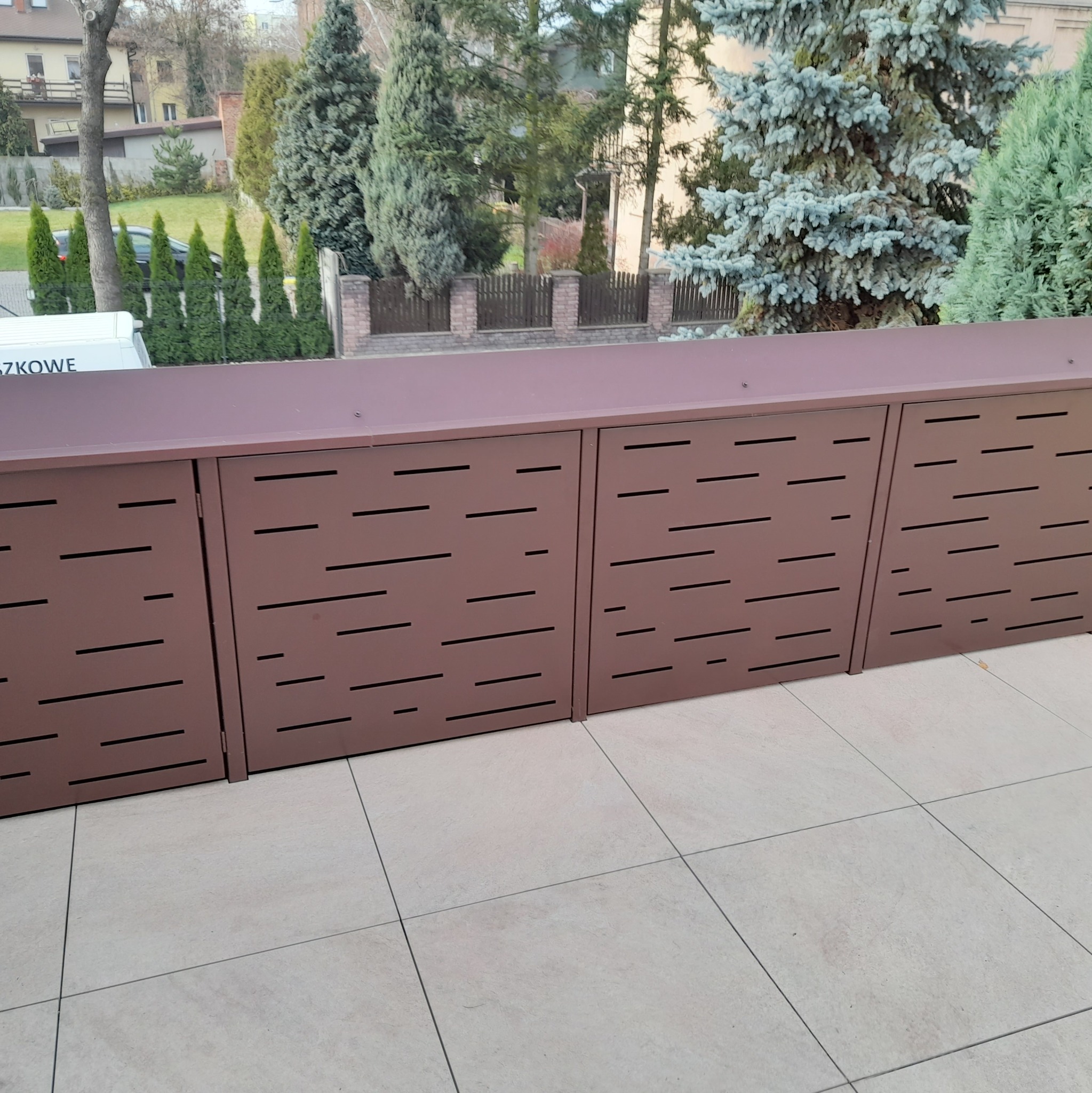 Nowoczesna, brązowa osłona balkonowa z poziomymi, prostokątnymi wycięciami, zamontowana na balkonie z jasnymi płytkami, z widokiem na drzewa i zabudowę mieszkalną w tle.