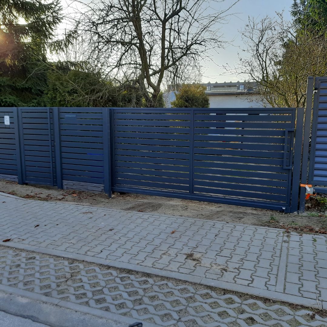 Nowoczesna, przesuwna brama wjazdowa z poziomych paneli, pomalowana na ciemny kolor, z widocznymi elementami montażowymi i kostką brukową przed posesją.