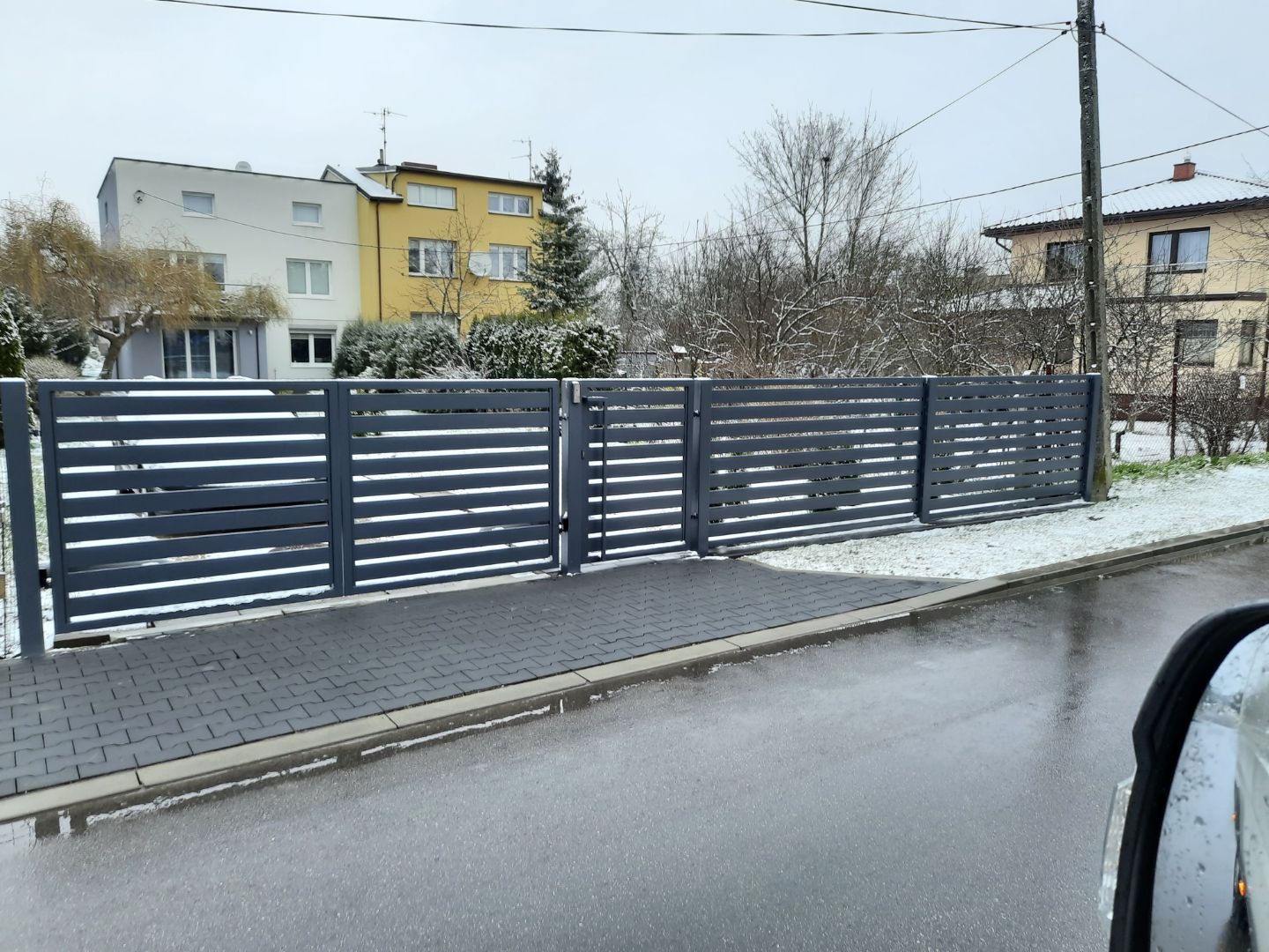 Nowoczesne, poziome, szare ogrodzenie panelowe z bramą wjazdową na tle dwukondygnacyjnego domu w zimowej scenerii.