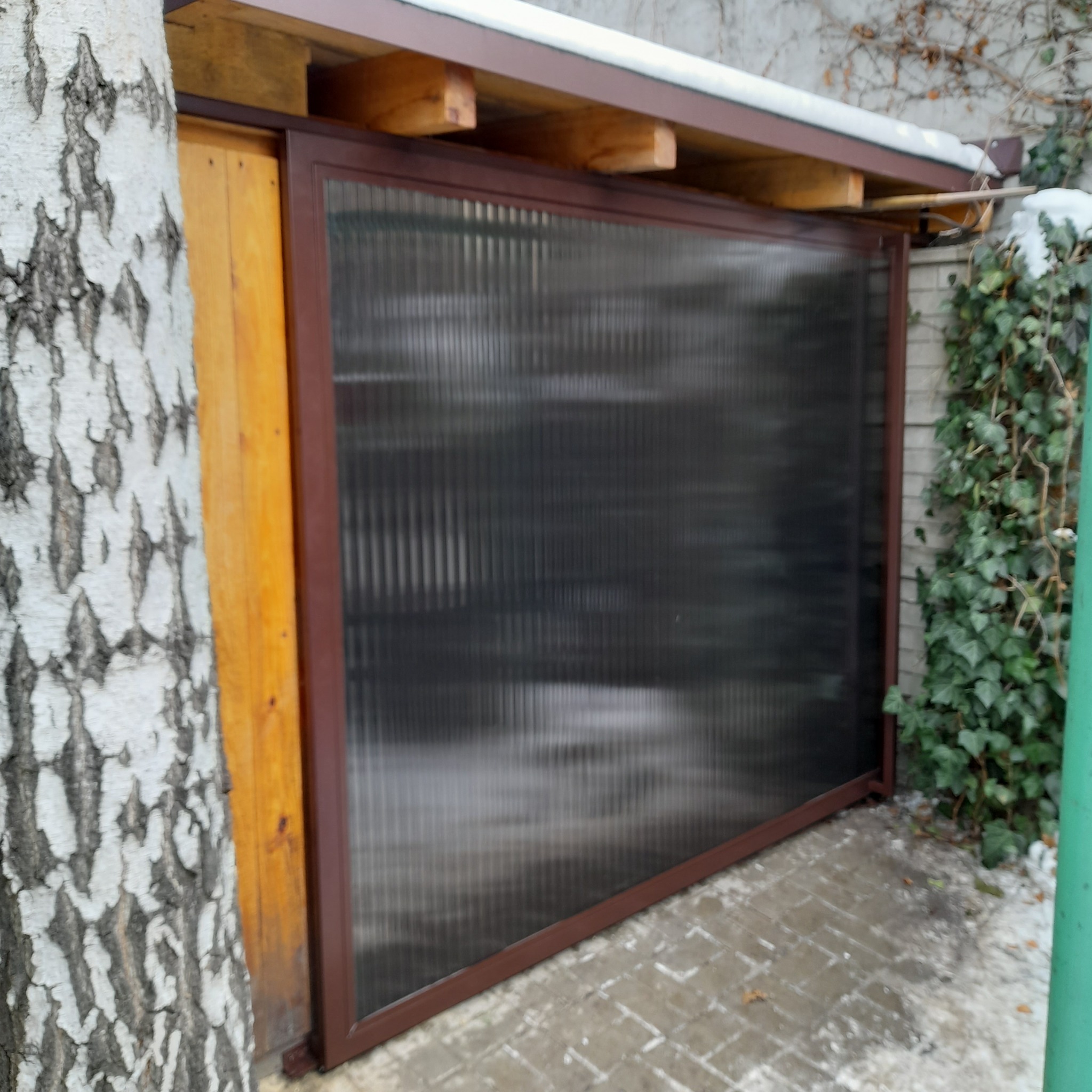 Przesuwna brama z brązową ramą i półprzezroczystym wypełnieniem, zamontowana obok drewnianego słupka i pnia brzozy, z fragmentami bluszczu i śniegu w tle.