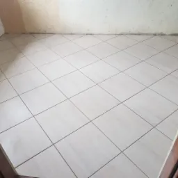 Jasne płytki podłogowe ułożone ukośnie w małym pomieszczeniu, widoczne surowe ściany w trakcie remontu i fragment drewnianej ościeżnicy drzwi.