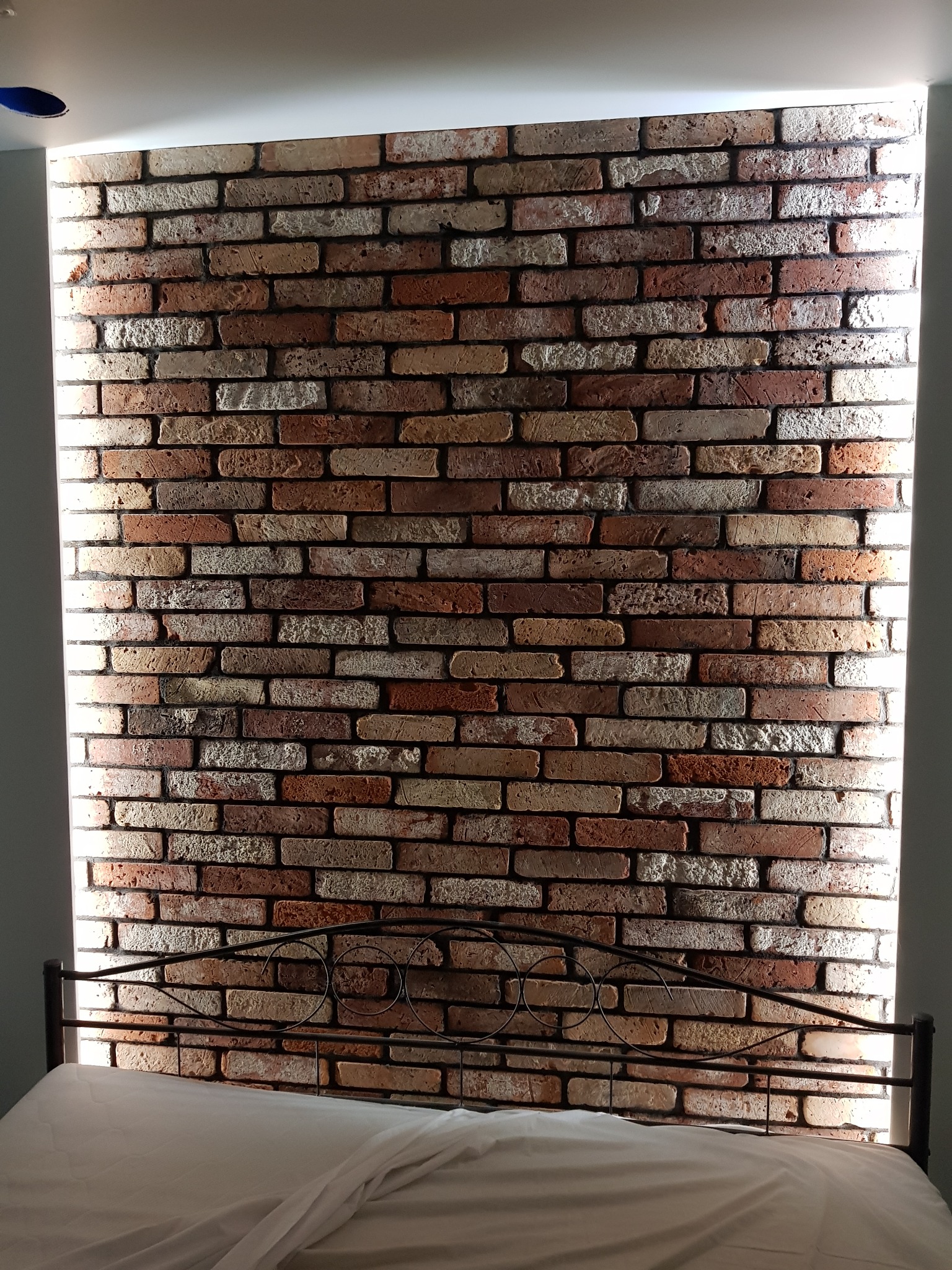 Ściana z dekoracyjnej cegły w sypialni, podświetlona od góry, widoczny fragment metalowego zagłówka łóżka