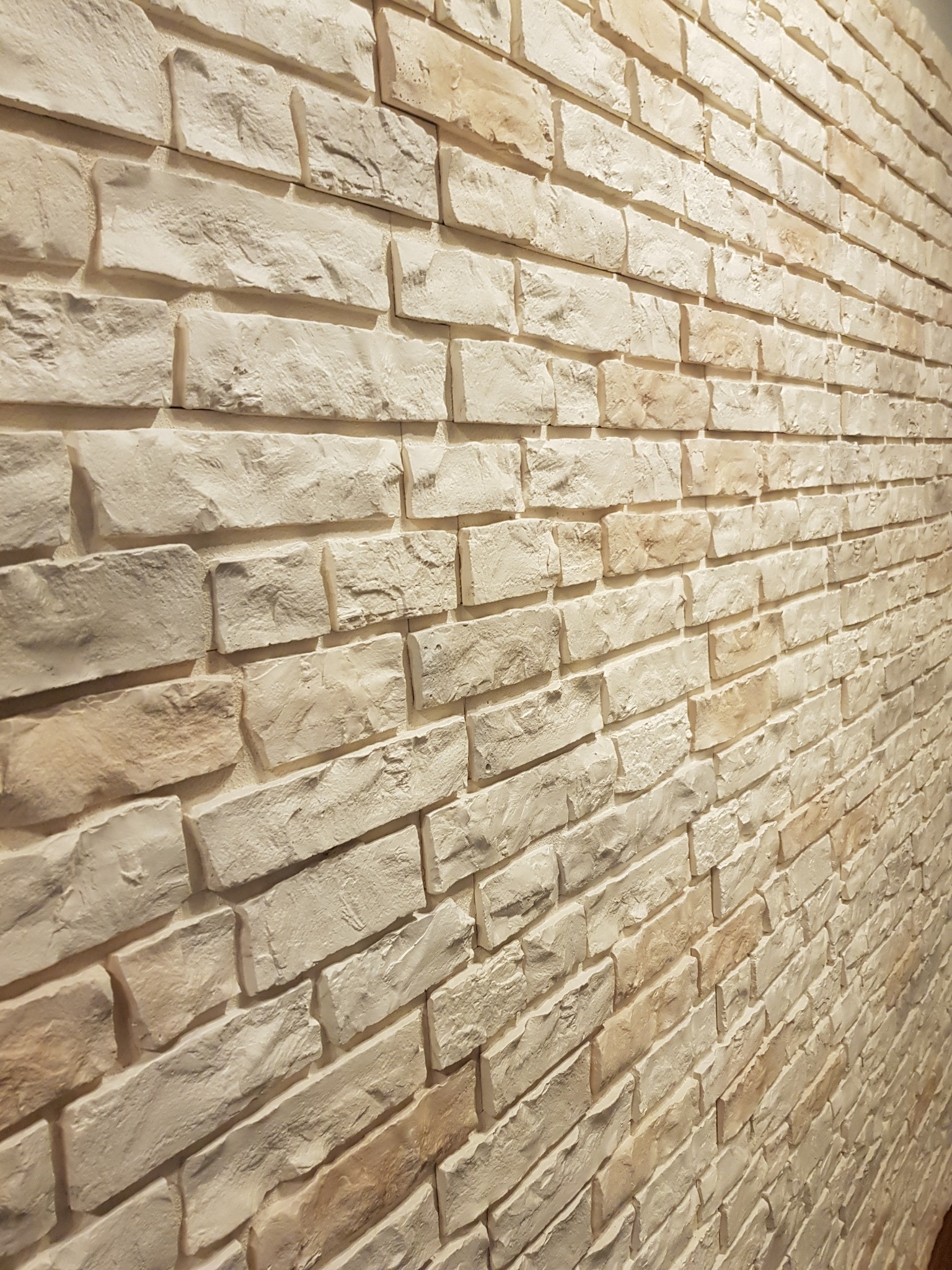 Ściana wyłożona dekoracyjną, beżowo-białą cegłą imitującą kamień naturalny, ułożona pod kątem, z widoczną strukturą i fakturą materiału.